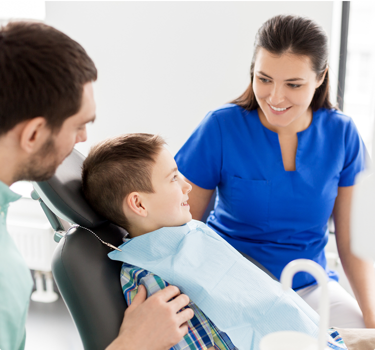 Young boy smiling in dental chair with parent beside him and hygienist talking before exam.