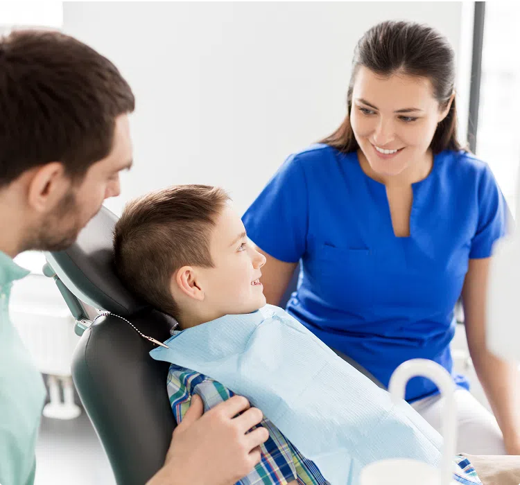 image (6) copy Young boy smiling in dental chair with parent beside him and hygienist talking before exam.