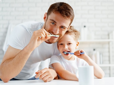 img copy 2 Father and young son smiling while brushing their teeth together in a bright bathroom.