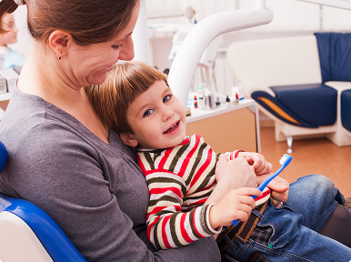 img copy Smiling toddler holding a toothbrush while sitting on mother’s lap at a dental clinic.