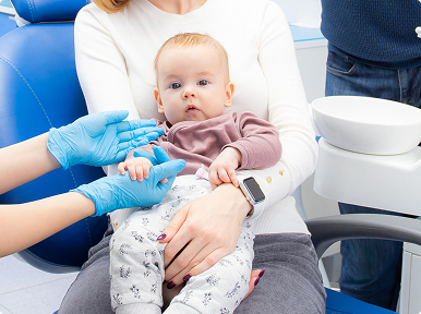 Infant sitting on mother’s lap during a first dental visit, with gloved hands reaching gently.