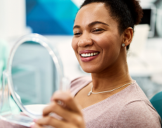 Woman smiling while looking at her reflection in a handheld mirror at a dental clinic.