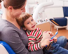 Smiling young boy holding a toothbrush while sitting on his mother’s lap at a dental clinic.