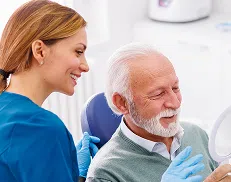 Elderly man smiling at his reflection in a mirror while a dental professional looks on.