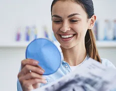 Woman smiling while looking at her reflection in a handheld mirror and holding dental X-rays.