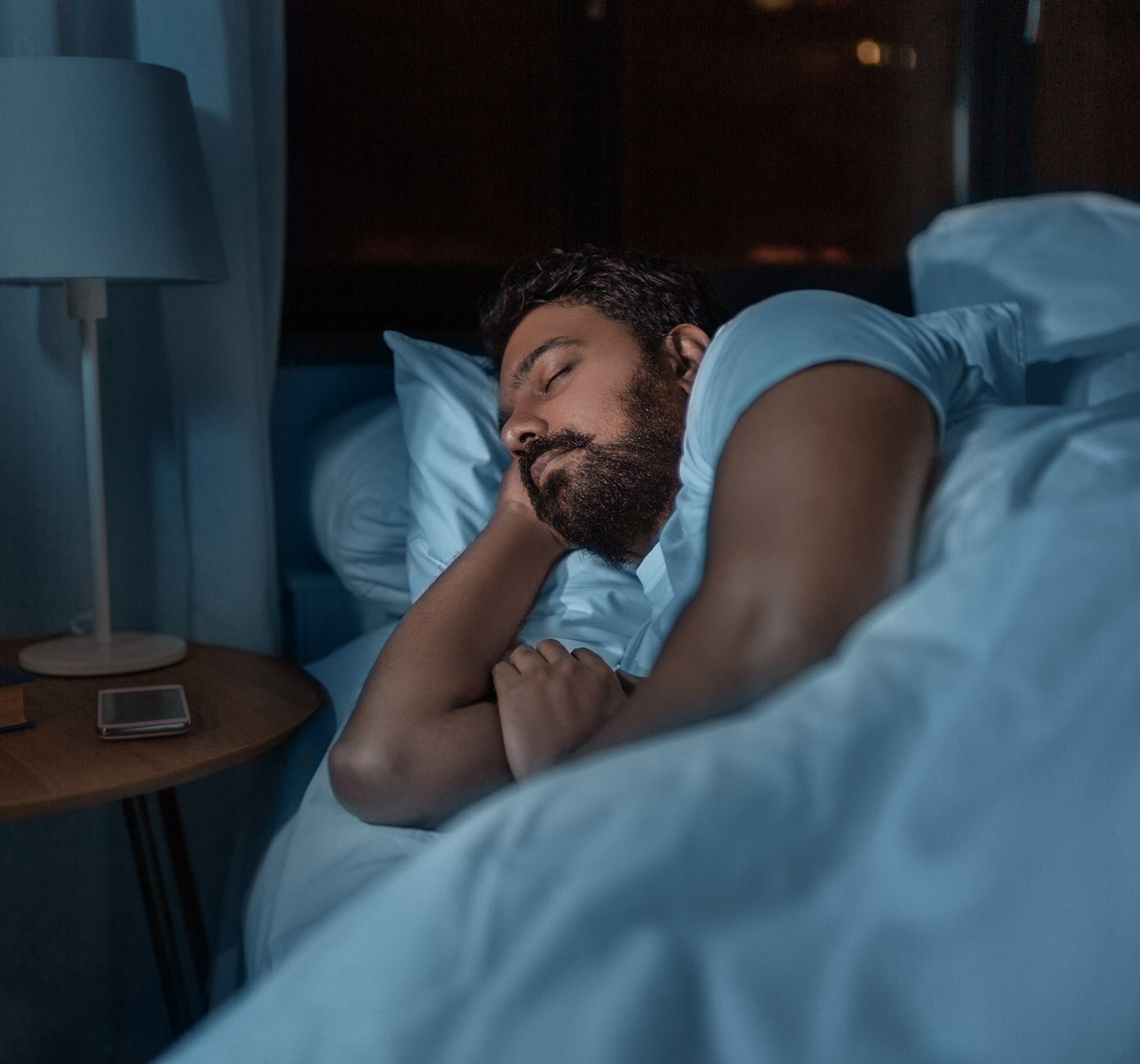 Man peacefully sleeping in bed at night, with a phone and lamp on the bedside table.