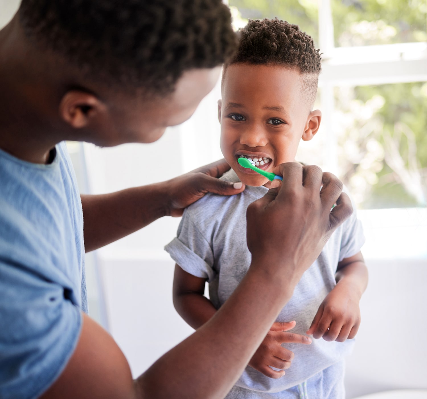 FSC-generaluse-image2-min Parent helping young child brush teeth with a green toothbrush in a bright bathroom.