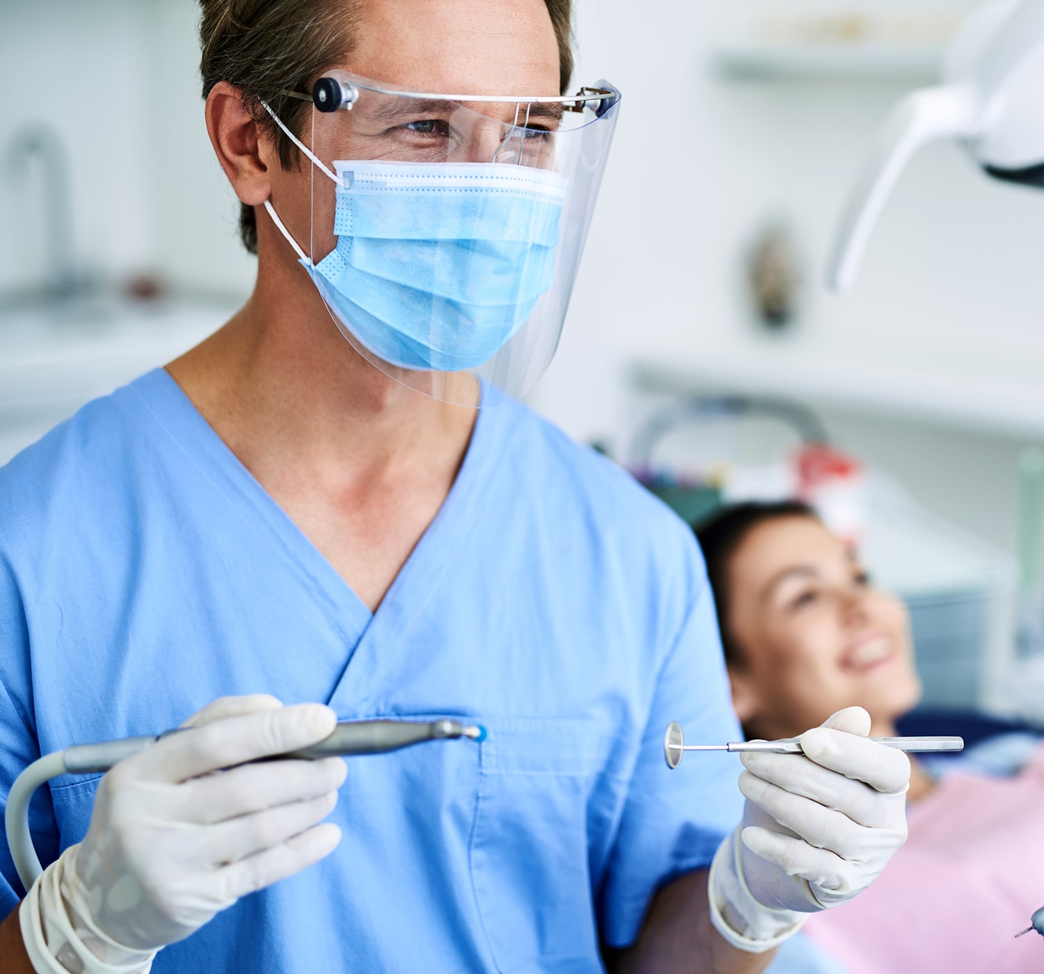 Dentist in protective gear holding tools, preparing to treat a smiling patient in a dental clinic.