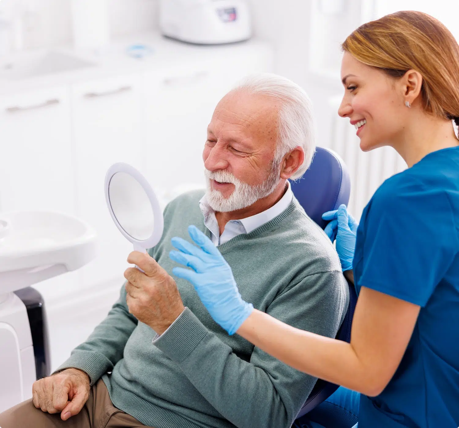 Older man smiling while looking in a hand mirror, assisted by a dental professional in a clinic.