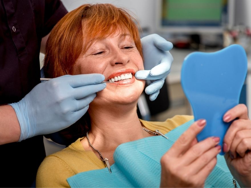 Restorative bottom CTA image 2 Middle-aged woman with red hair looking at smile in mirror during dental appointment