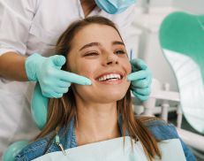 A young woman is sitting in a dental exam chair. The hygienist is using her pointer fingers to trace a smile at the corners of the woman's mouth