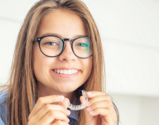 A young girl with brown hair smiling and wearing glasses holding her Invisalign clear aligner trays.
