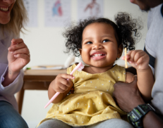 A happy smiling baby with curly hair at the dentist holding a tooth brush